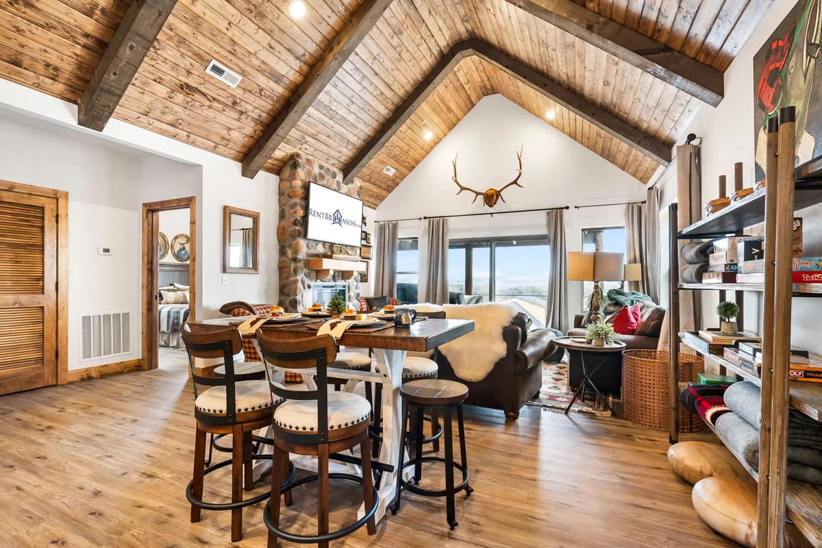 A spacious, open-concept living and dining area with a high vaulted wooden ceiling, a stunning stone fireplace, large windows that let in natural light and offer scenic views, and a dining table set for a gathering. The space is arranged for family gatherings and cozy reading, blending comfort and rustic elegance.