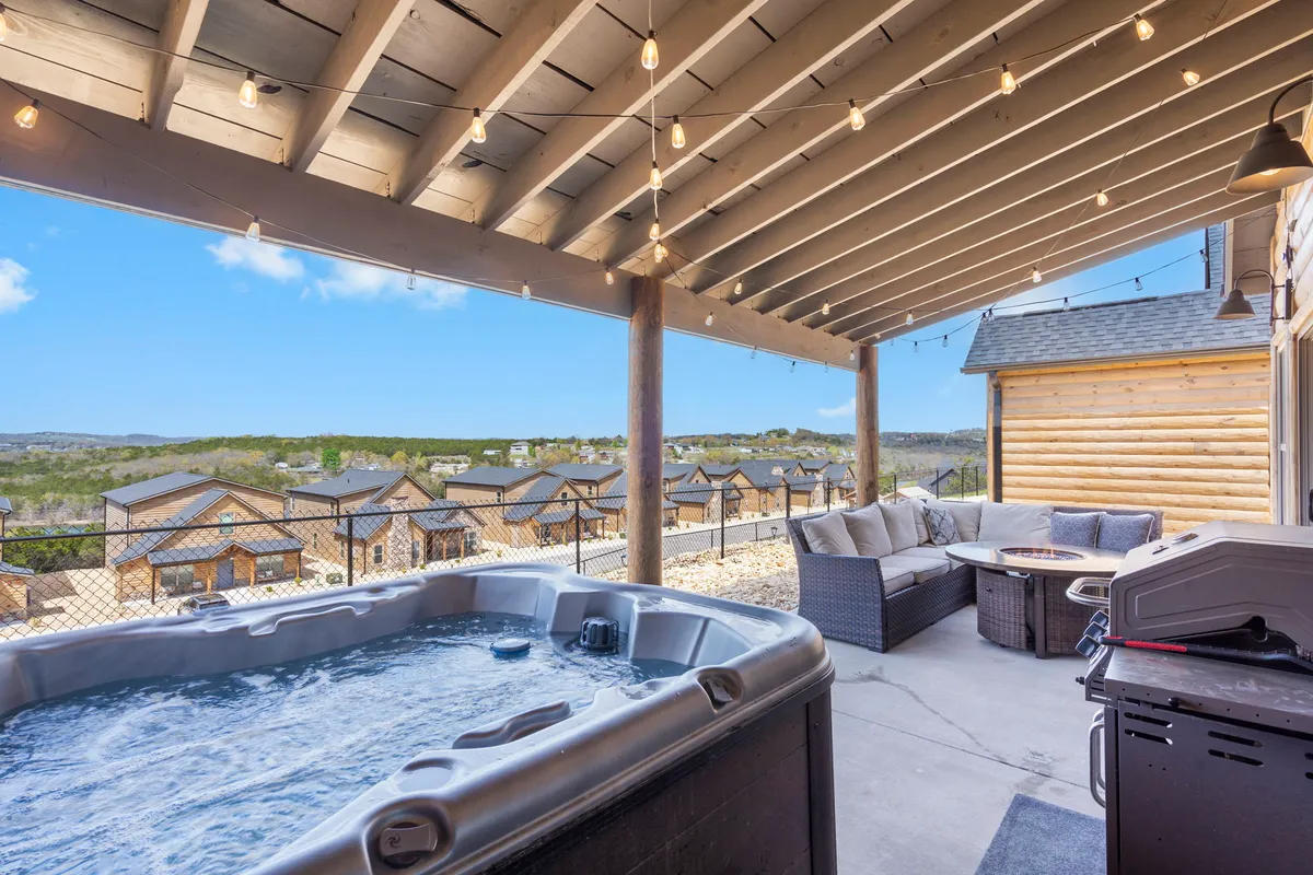 A tranquil patio space at dusk features a hot tub, comfortable outdoor seating, hanging string lights, and a landscape view that hints at the nearby lake. The ambiance is both relaxing and inviting, ideal for sunset drinks or stargazing evenings.