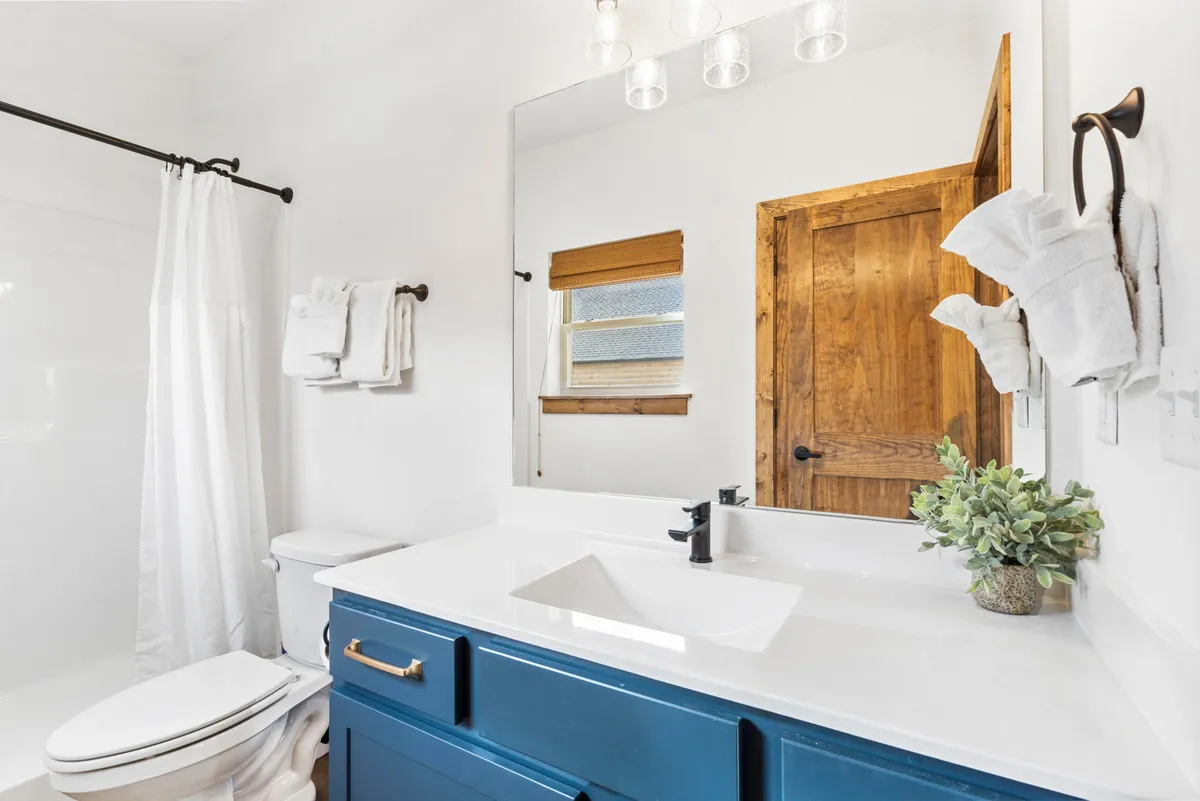 A bright, tranquil ensuite bathroom featuring a blue vanity, large mirror, white shower curtain, and green plant. The space is fresh, stylish, and designed for personal routines, contributing to an overall sense of comfort and privacy.