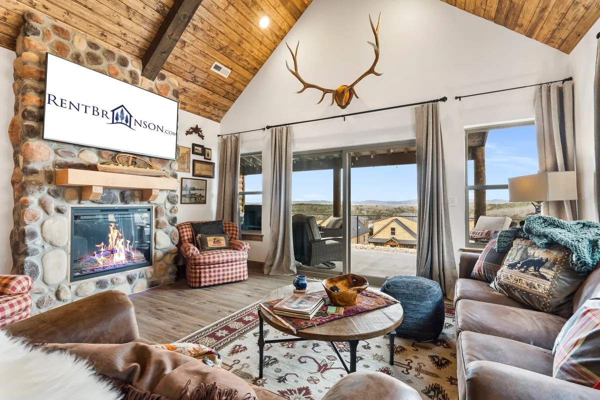 The image showcases a warm and inviting log cabin living room, featuring a crackling fireplace beneath rustic antlers, expansive windows framing Ozark views, and plush seating arranged perfectly for relaxation and connection.