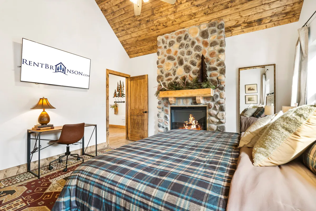 A cozy king bedroom featuring a plush bed with a plaid blanket, a warm stone fireplace, a TV mounted on the wall, and a wooden door. The atmosphere is comforting and inviting, perfect for unwinding and enjoying private time. The room exudes a sense of sanctuary and comfort, highlighting the in-room fireplace.