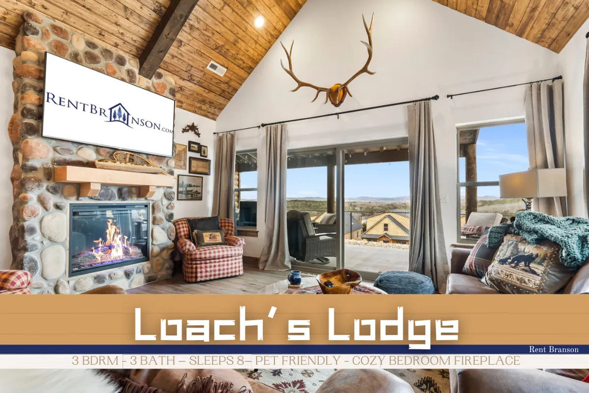 This interior scene features a grand stone fireplace with a roaring fire, large windows that frame a beautiful outdoor view, rustic wooden beams overhead, and antler decor. Plush seating is arranged for comfort and socializing, suggesting a cozy and inviting atmosphere perfect for relaxing indoors.
