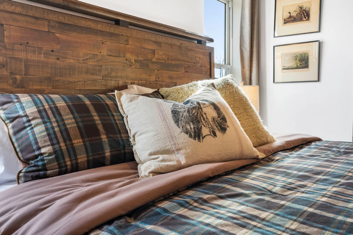 A tranquil bedroom adorned with plaid bedding, textured pillows, a wooden headboard, and framed artwork. Gentle sunlight filters through the window, creating an inviting and restful space ideal for relaxation and restful sleep.