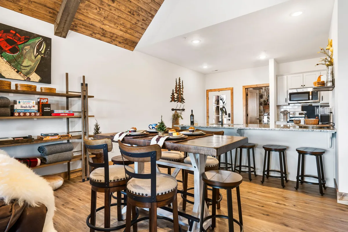 A warm, welcoming dining area featuring a wooden dining table, open shelving with board games and decorative items, and a nearby kitchen area. The scene suggests a lively space for family meals, game nights, and shared cooking experiences.